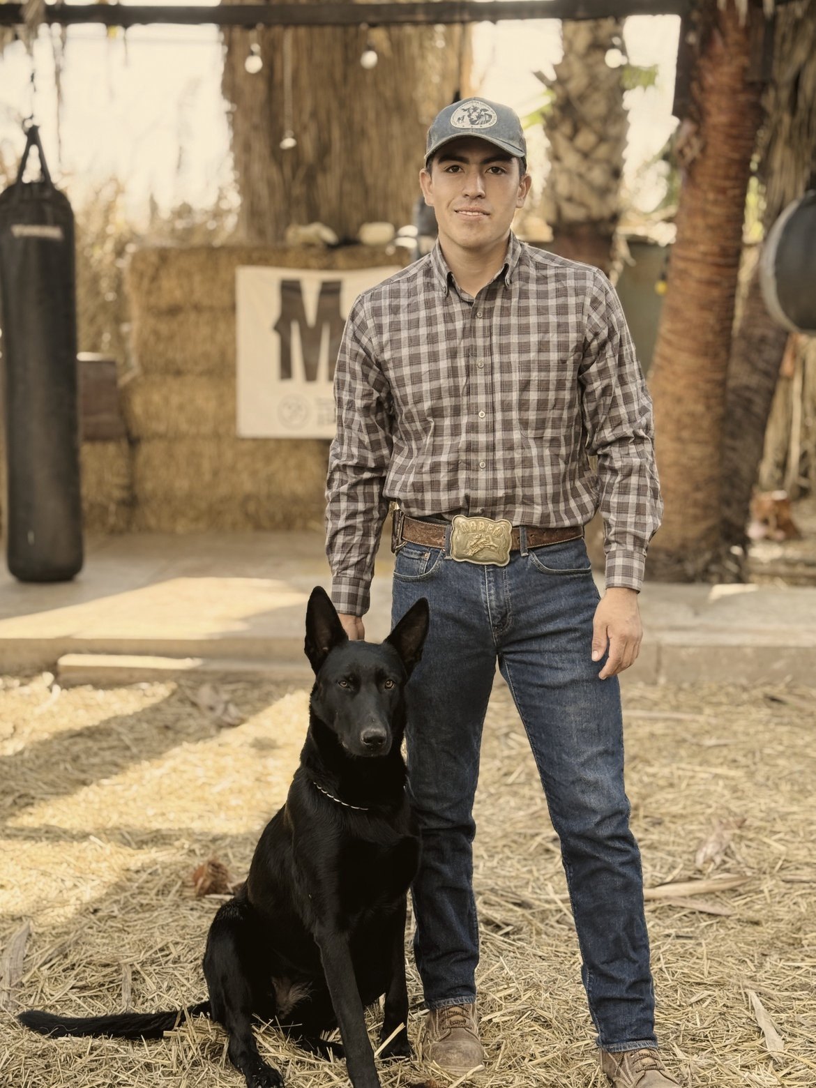 Abel Núñez Nuño, in a plaid shirt, cap, jeans and a rodeo belt buckle, stands on straw beside a black shepherd dog at the Mud & Lotus Baja ranch