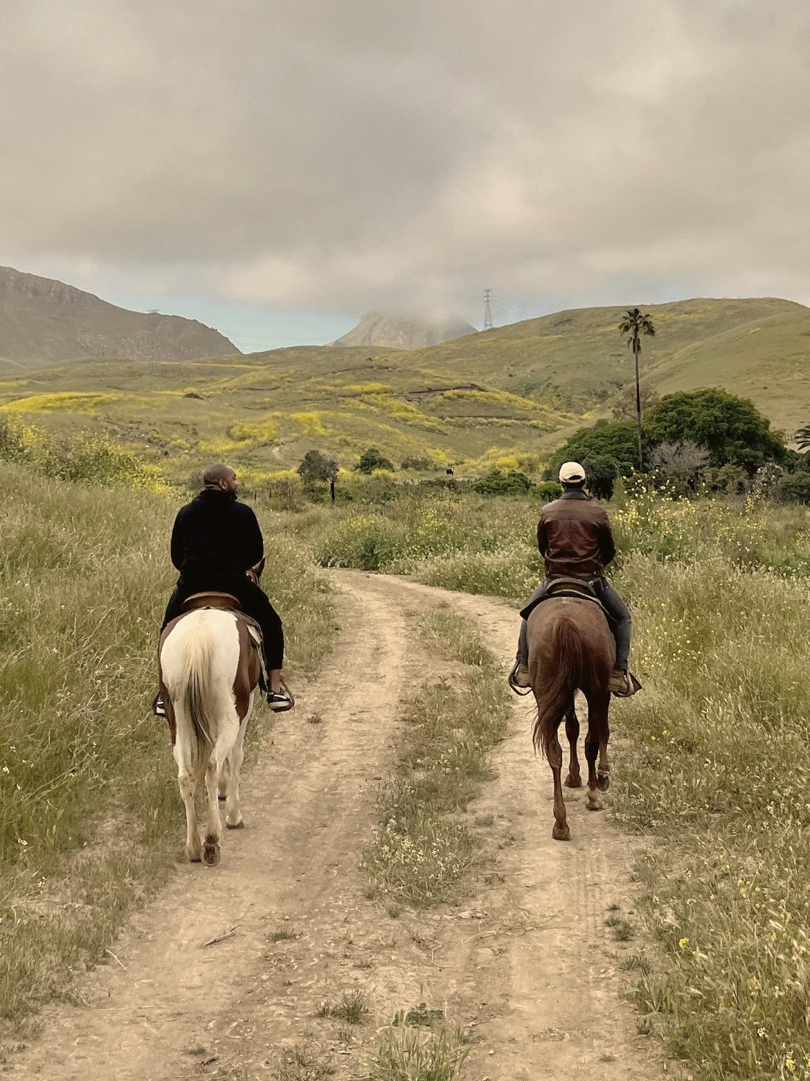Two riders follow a dirt trail through green hills under clouds.