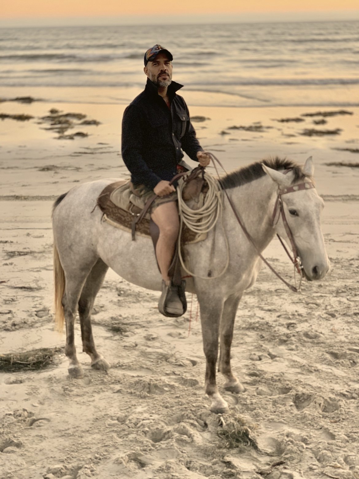 Anas “Coach” Canon, in a dark jacket and cap, sits on a gray horse with a coiled rope at the saddle, on the beach at sunset