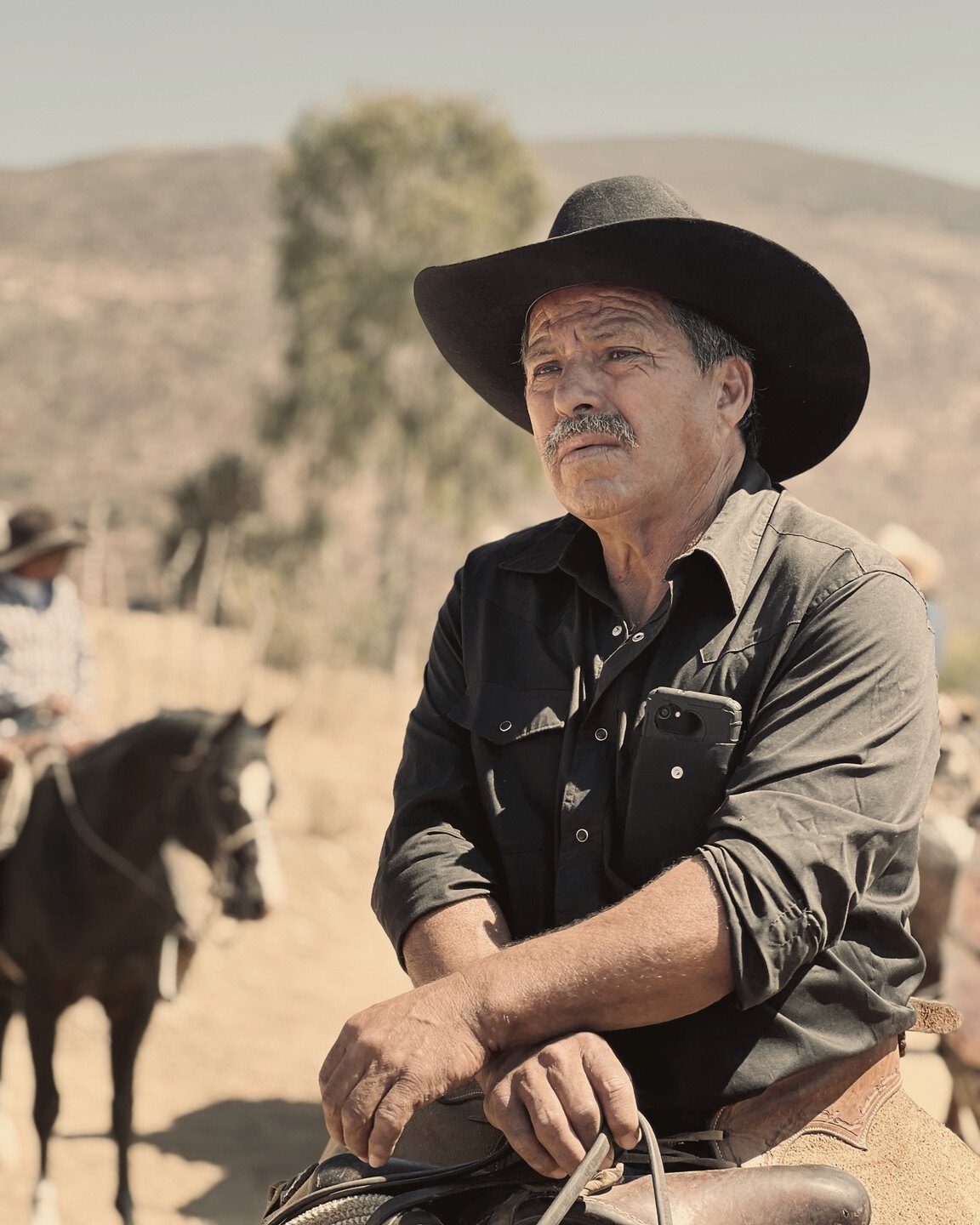 A mustached rider in a black hat and shirt rests his arms across a saddle, with another horse behind him
