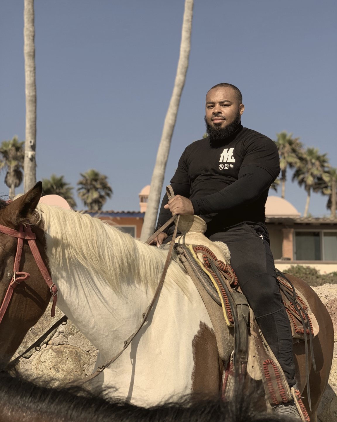 A bearded rider in a black Mud & Lotus T-shirt sits on a brown-and-white paint horse under palm trees.